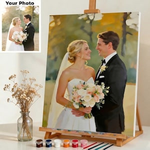 May include: A wedding portrait painting on an easel. The painting depicts a smiling couple, the bride in a white strapless gown holding a bouquet. The groom wears a black tuxedo. The painting is on a wooden easel with paint and brushes.