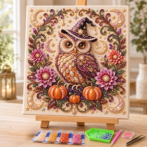 May include: A diamond painting kit featuring a square canvas with a central owl wearing a purple witch hat, surrounded by pink flowers, orange pumpkins, and crescent moons. The design is embellished with colorful beads and gold accents.