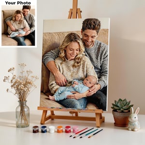 May include: A custom canvas print on a wooden easel displaying a family portrait of a woman, a man, and a baby. The set includes small paint pots and brushes for painting by numbers. A glass vase with dried flowers and a ceramic rabbit are nearby.