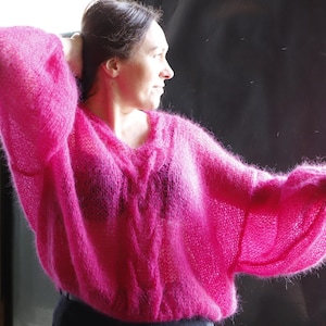 May include: A woman wearing a bright pink fuzzy sweater with a cable knit design. The sweater has long sleeves and a loose fit.