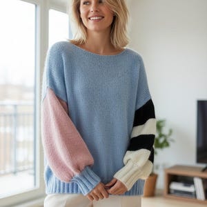 May include: A light blue sweater with color block sleeves. One sleeve is pink, and the other has black and white stripes. The sweater has a round neckline and is made of a soft, knitted material. The model is wearing white pants.