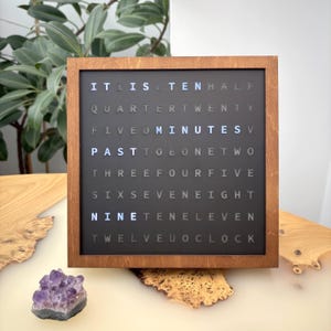Peut inclure: Une horloge carrée avec un cadre en bois et un cadran gris foncé. L'heure est affichée en mots, avec des lettres éclairées. Le cadre est brun chaud. Un cristal d'améthyste violet est en dessous.