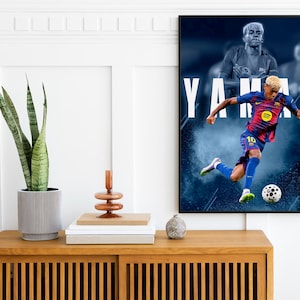 K&ouml;nnte beinhalten: Ein gerahmtes Poster mit einem Fu&szlig;ballspieler in blau-roter Uniform, der einen Ball tritt. Der Name des Spielers, YAMAL, ist in gro&szlig;en wei&szlig;en Buchstaben dargestellt. Das Poster h&auml;ngt &uuml;ber einem Holzschrank mit einer Pflanze und Dekoration.