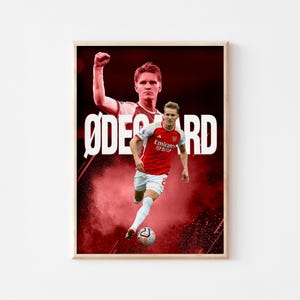 May include: A framed poster featuring a soccer player in a red and white jersey, with the name "ØDEGAARD" in large white letters. The background is a red and black abstract design with a soccer ball at the player's feet.