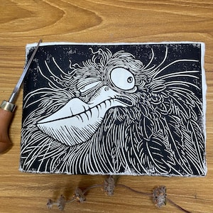 May include: Black and white linocut print of a whimsical bird with a large beak and expressive eye. The print is on a white rectangular paper. A carving tool with a wooden handle is visible on the left side.