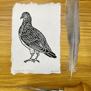 May include: A black and white linocut print of a pigeon titled "Judgmental Pigeon" on textured white paper. A gray feather and a carving tool are next to the print. The print is signed "ks 2025".