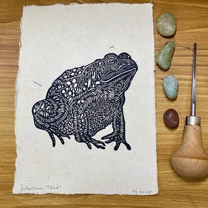 Peut inclure: Une linogravure en noir et blanc d'un crapaud de l'Ouest sur papier texturé. L'œuvre présente des détails complexes et est signée "KS 2025". À côté de l'impression se trouvent de petites pierres et un outil de sculpture.
