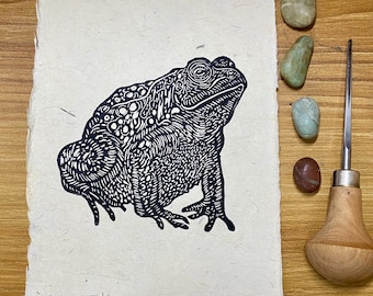 May include: A black and white linocut print of a Western Toad on textured paper. The artwork features intricate details and is signed "KS 2025". Beside the print are small stones and a carving tool.