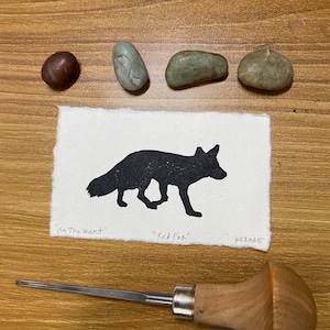 May include: A black silhouette of a fox on a white paper with the text "on the Hunt" and "Red Fox". A wood carving tool and several small stones are also present.
