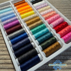 May include: A white plastic organiser filled with spools of colourful thread. The thread colours include silver, orange, yellow, gold, teal, blue, and pink. The spools are neatly arranged in rows.
