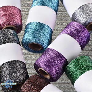 Puede incluir: Una colección de coloridas bobinas de hilo metálico. Las bobinas están envueltas con etiquetas blancas y vienen en varios colores, incluyendo rosa, turquesa, plata, morado, verde y marrón. El hilo tiene una calidad brillante y reflectante.