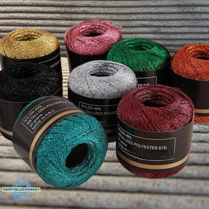 Op de afbeelding: Een verzameling kleurrijke metallic garenspoelen in goud, roze, groen, zilver, oranje, zwart, blauwgroen en bordeauxrood. Elke spoel is gelabeld met details, waaronder de materiaalsamenstelling en het gewicht van 20 gram.