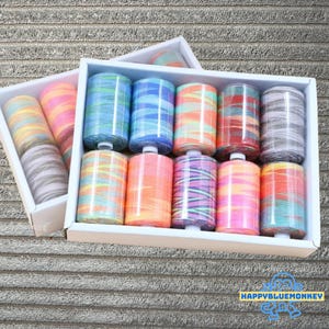 Könnte beinhalten: Ein Set bunter Garnrollen in einer weißen Box. Die Rollen sind in Reihen angeordnet und weisen eine Vielzahl von Farben auf, darunter Blau, Orange, Rosa und Grau. Der Faden ist in einem gestreiften Muster um die Rollen gewickelt. Die Box steht auf einer strukturierten Oberfläche.