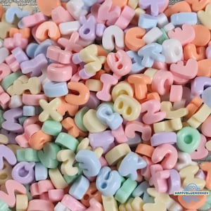 Op de afbeelding: Een grote verzameling kleine, pastelkleurige alfabetletterkralen in tinten roze, blauw, geel, oranje en groen. Elke kraal heeft een afgerond, dik lettertype met een gat in het midden om te rijgen. De tekst HAPPYBLUEMONKEY is zichtbaar.