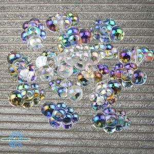 Può includere: Una collezione di perline in vetro trasparente iridescente a forma di grappoli di bolle. Ogni perlina presenta una finitura riflettente multicolore con tonalità blu, viola e oro. Le perline hanno un foro centrale e una superficie liscia.