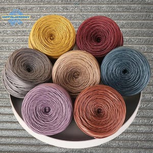 May include: A selection of colourful yarn balls in shades of yellow, burgundy, grey, tan, blue, lavender, and rust. The yarn is arranged in a circular pattern within a white, shallow dish. The image includes the text "HAPPYBLUEMONKEY".