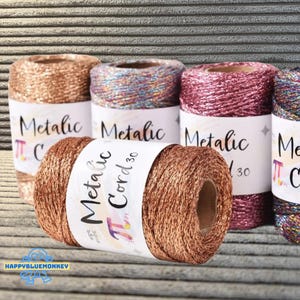 May include: A selection of metallic cord spools in shades of bronze, pink, and iridescent colours. Each spool features a white label with the text "Metallic Cord 30". The cord is presented against a textured grey backdrop.