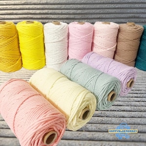 May include: A selection of colourful macrame cord spools in shades of yellow, white, pink, beige, and teal. The spools are arranged in rows, showcasing the variety of colours available for crafting projects. The Happy Blue Monkey logo is visible.