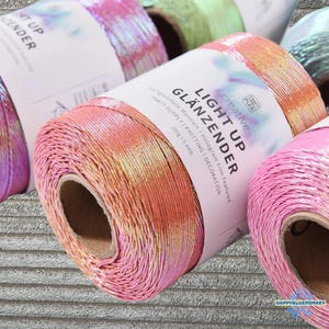 May include: A spool of iridescent peach and pink yarn with a white paper label reading LIGHT UP GLÄNZENDER, creative CRAFT PLEX, hologram film featured, party supply, knitting, decoration, and 200g / 0.44lb, surrounded by other colorful yarn spools.