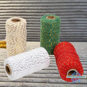 May include: Four spools of twine arranged in a group. One spool is cream with gold metallic thread, one is green with gold metallic thread, one is white with black thread, and one is red with gold metallic thread. Each spool has a cardboard core.