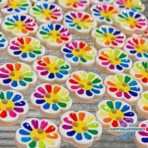 May include: A collection of flower-shaped buttons with a white base and colorful rainbow petals. Each button has a yellow center and a small hole for sewing. The buttons are arranged on a textured gray surface. The text "HAPPYBLUEMONKEY" is visible.