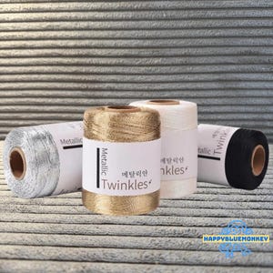 Könnte beinhalten: Vier Spulen Metallic-Garn in Silber, Gold, Weiß und Schwarz. Jede Spule hat ein Etikett mit dem Wort "Metallic" und dem Markennamen "Twinkles". Die Spulen sind auf einer strukturierten Oberfläche angeordnet.