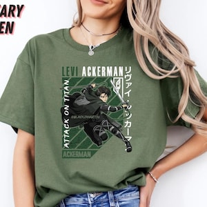 May include: Military green t-shirt featuring a graphic of Levi Ackerman from Attack on Titan. The design includes text in English and Japanese, with a dynamic illustration of the character in action. The shirt is a solid color.
