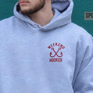 Könnte beinhalten: Ein sportgrauer Kapuzenpullover mit einer Kängurutasche auf der Vorderseite und einem gestickten Motiv auf der linken Brust. Das Design besteht aus zwei gekreuzten roten Angelhaken mit dem Text WEEKEND HOOKER in roten Großbuchstaben.
