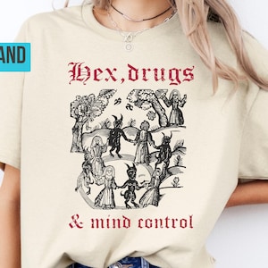 May include: A sand-coloured t-shirt with a black and white illustration of figures in a forest, and the red text "Hex, drugs & mind control" in gothic lettering. The shirt is a casual, short-sleeved style.