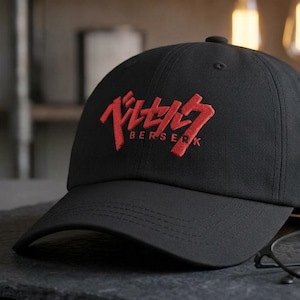 May include: Black baseball cap featuring the red embroidered word "BERSERK". The cap has a curved peak and a structured fit. The hat is displayed on a dark grey surface.