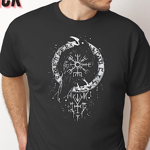 K&ouml;nnte beinhalten: Schwarzes T-Shirt mit kurzen &Auml;rmeln und einem wei&szlig;en Raben- und Runen-Motiv. Das Wort "BLACK" ist in Rosa oben links aufgedruckt. Das Design ist kreisf&ouml;rmig.