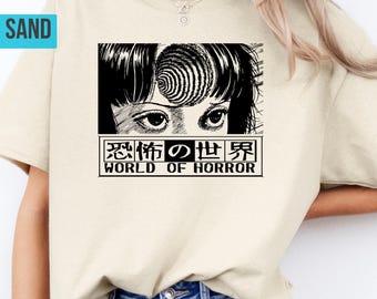 Op de afbeelding: Zandkleurig T-shirt met een zwart-witte manga-stijl illustratie en de tekst "WORLD OF HORROR" in het Engels en Japans. Het T-shirt wordt gedragen met een blauwe spijkerbroek.