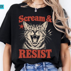 Könnte beinhalten: Ein schwarzes Kurzarm-T-Shirt mit einer Grafik einer gähnenden getigerten Katze und dem Text "Scream & Resist" in roter Schrift. Das Design enthält einen kleinen roten Stern und Blitzakzente, die auf die Vorderseite des Baumwollstoffs gedruckt sind.