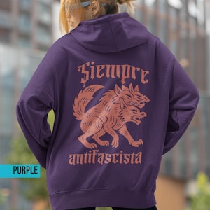 Könnte beinhalten: Ein lila Kapuzenpullover mit einem zweiköpfigen Wolf-Motiv in Rostfarbe auf dem Rücken. Die Wörter "Siempre" und "antifascista" befinden sich über und unter der Wolf-Illustration. Das Wort "PURPLE" steht in einem blauen Rechteck unten links.