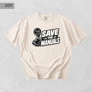 Może przedstawiać: Kremowa koszulka z czarno-białą grafiką dźwigni zmiany biegów i napisem "SAVE THE MANUALS". Koszulka z krótkim rękawem i okrągłym dekoltem ma centralnie umieszczony wzór. Koszulka wykonana jest z miękkiego materiału.