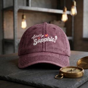 May include: A maroon baseball cap with a distressed finish featuring the embroidered white text "Totally Sapphic!" and two small pink and yellow flower icons on the front panel. The cap has a curved brim and a structured six-panel design.