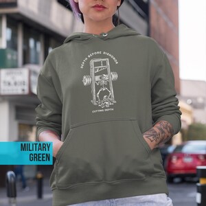 Puede incluir: Sudadera con capucha verde militar con bolsillo delantero y capucha con cord&oacute;n. El gr&aacute;fico en la parte delantera presenta un esqueleto en una guillotina con las palabras "DEPTH BEFORE DISHONOR" y "CUTTING DEPTH".