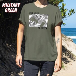 May include: Military green t-shirt with the text "EAT YOUR PROTEIN" and a black and white graphic. The graphic depicts a manga-style illustration of a creature. The shirt is a solid color.