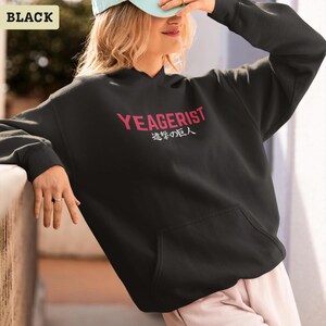 Puede incluir: Una sudadera negra con capucha y mangas largas que presenta el texto YEAGERIST en letras may&uacute;sculas rojas en el pecho, con caracteres japoneses impresos en blanco justo debajo. La prenda incluye un bolsillo tipo canguro y pu&ntilde;os acanalados.