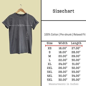 K&ouml;nnte beinhalten: Ein graues T-Shirt h&auml;ngt an einem Holzb&uuml;gel, daneben eine Gr&ouml;&szlig;entabelle. Die Tabelle zeigt Ma&szlig;e in Zoll f&uuml;r die Gr&ouml;&szlig;en XS bis 5XL, mit Breiten von 40.64 cm bis 81.28 cm und L&auml;ngen von 68.58 cm bis 88.90 cm.