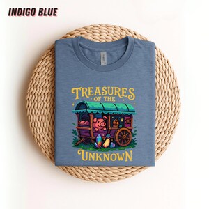 May include: Indigo blue t-shirt featuring a whimsical illustration. The design includes a pig in a caravan with the text "Treasures of the Unknown" in yellow and green. The shirt is displayed on a woven, circular mat.