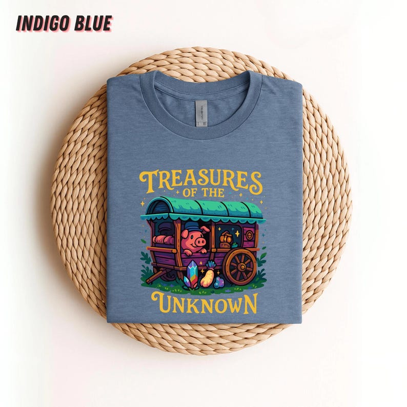 May include: Indigo blue t-shirt featuring a whimsical illustration. The design includes a pig in a caravan with the text "Treasures of the Unknown" in yellow and green. The shirt is displayed on a woven, circular mat.