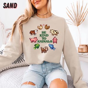 May include: Sand-colored sweatshirt with the text "BE KIND TO ANIMALS" surrounded by pixelated farm animals. The sweatshirt has a crew neck and long sleeves. The word "SAND" is printed in a bubble font in the upper left corner.