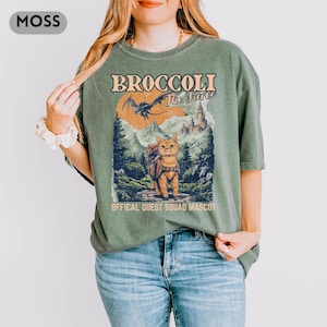 May include: Moss green t-shirt with a graphic featuring a cat with a backpack, a dragon, and a castle. The text reads "BROCCOLI The Fierce" and "OFFICAL QUEST SQUAD MASCOT". The shirt is a relaxed fit.