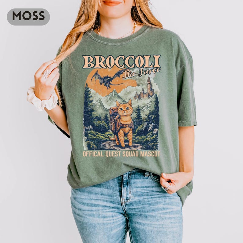 May include: Moss green t-shirt with a graphic featuring a cat with a backpack, a dragon, and a castle. The text reads "BROCCOLI The Fierce" and "OFFICAL QUEST SQUAD MASCOT". The shirt is a relaxed fit.