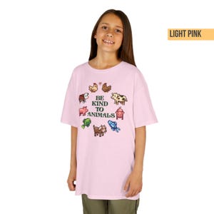 May include: Light pink t-shirt with the text "BE KIND TO ANIMALS" and pixelated animal graphics. The design includes a cow, pig, fish, goat, and other farm animals. The shirt is a soft, comfortable material.