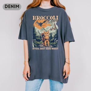 May include: Denim blue t-shirt featuring a graphic of a cat with a backpack in a forest scene, with text that reads "BROCCOLI The Fierce" and "OFFICAL QUEST SQUAD MASCOT". The shirt has a crew neck and short sleeves.