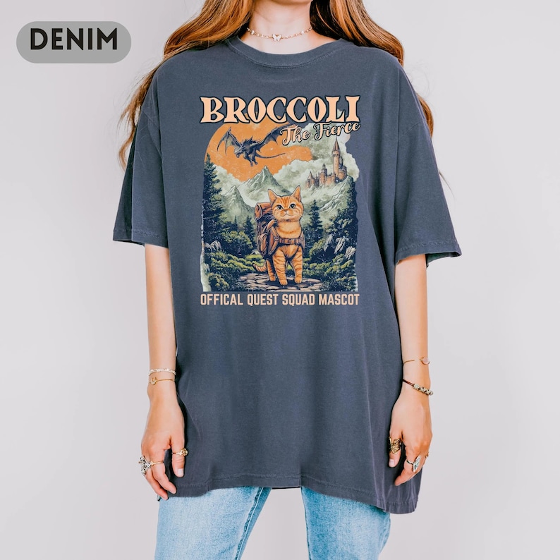 May include: Denim blue t-shirt featuring a graphic of a cat with a backpack in a forest scene, with text that reads "BROCCOLI The Fierce" and "OFFICAL QUEST SQUAD MASCOT". The shirt has a crew neck and short sleeves.