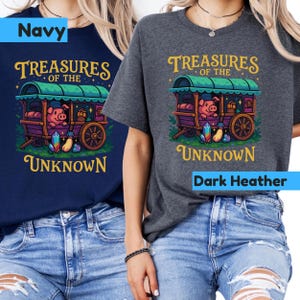 May include: Navy and dark heather t-shirts featuring a whimsical illustration. The design includes a pig in a wagon with the text "Treasures of the Unknown" in gold lettering. The wagon is purple and green, with colorful gems.