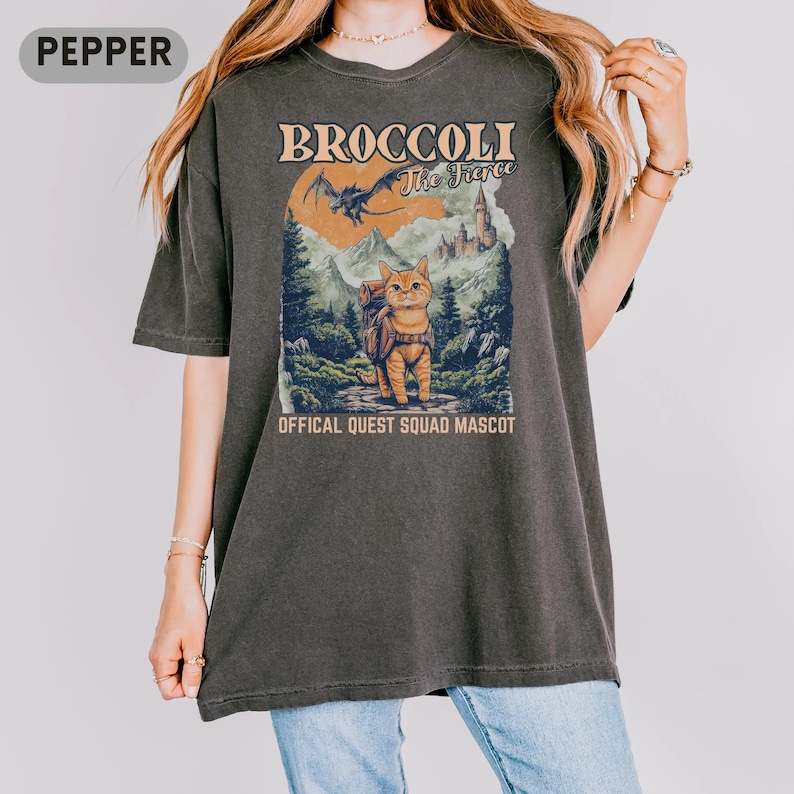 May include: Dark gray t-shirt featuring a cartoon cat with a backpack, set against a mountain and castle backdrop. The shirt reads "Broccoli The Fierce" and "Official Quest Squad Mascot". The design includes a dragon and a sunset.